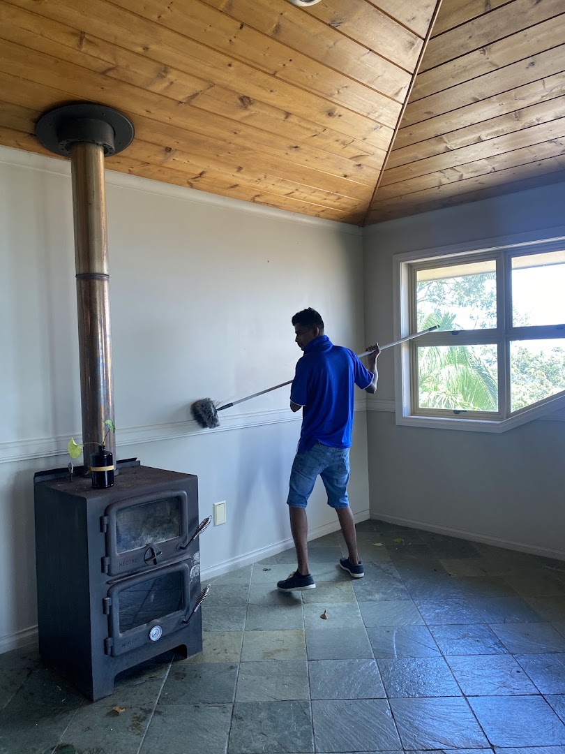 A cleaner in a blue top reaching the top of a wall with a long-handled duster beside a wood heater