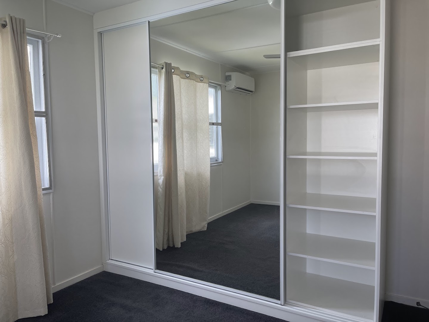 A bedroom with a mirrored sliding robe and open shelving beside a curtained window
